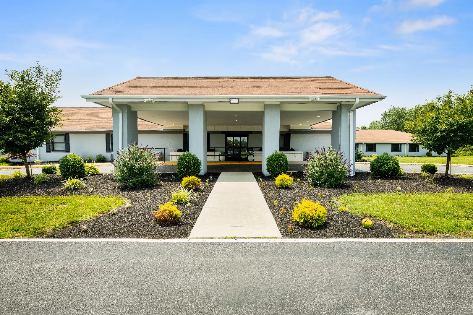 Front entrance of a single-story senior living facility with a covered portico, walkway, and landscaped flowerbeds.