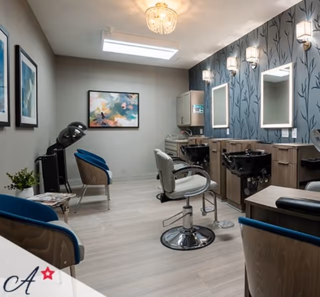 Interior view of a salon area in an assisted living facility featuring salon chairs, hair washing stations with sinks, illuminated mirrors on a patterned blue wall, a hair dryer, framed artwork, and modern lighting fixtures.