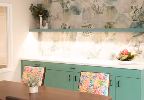 A dining area with a wooden table, two colorful patterned chairs, green cabinets, a floating shelf and botanical wallpaper.