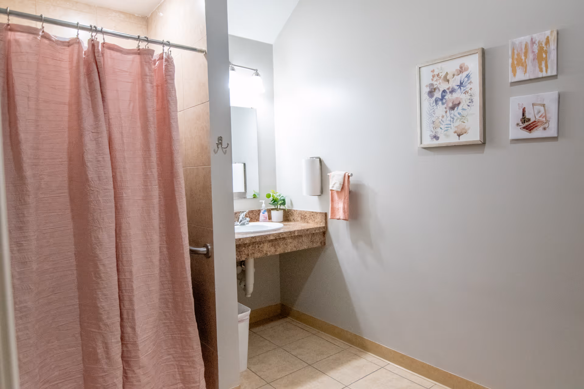 A bathroom with a pink shower curtain on a metal rod, a beige tiled shower wall, a granite countertop with a white sink, a mirror above the sink, a small potted plant, a soap dispenser, a towel rack with a pink towel and a white towel, and three framed artworks on a light gray wall.