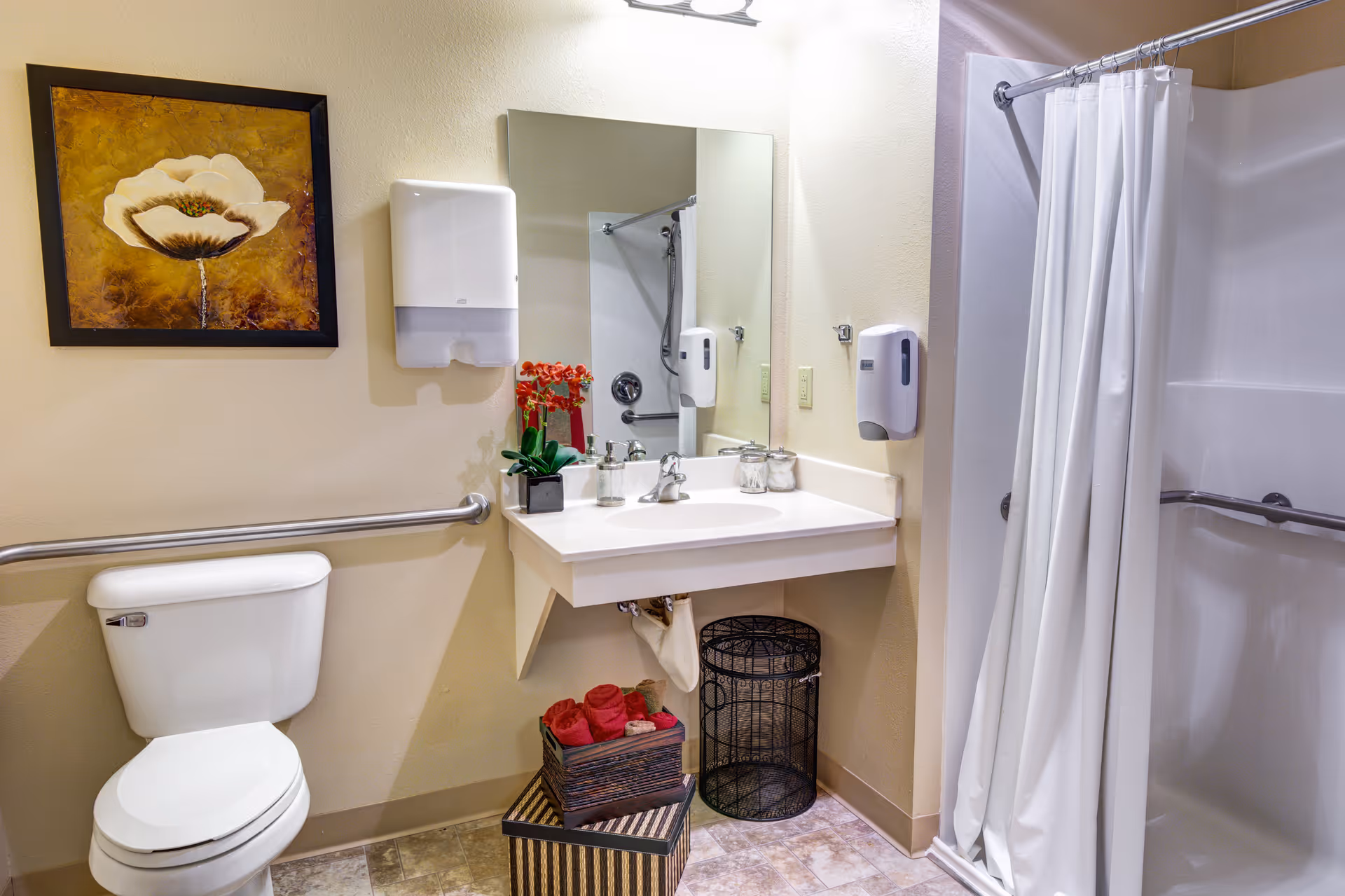 A clean and accessible bathroom featuring a white toilet with a grab bar, a wall-mounted sink with a large mirror above it, a soap dispenser, and a shower area with a white curtain. There is a framed flower painting on the wall and decorative red flowers on the sink. A basket with rolled red towels and a black wire wastebasket are placed under the sink.