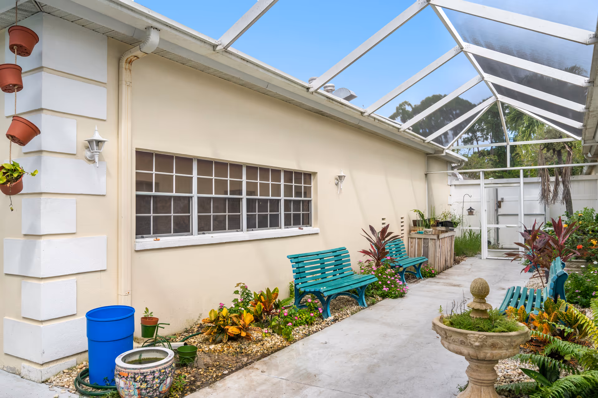 A covered outdoor patio area with a transparent roof, featuring green benches, potted plants, and a small garden with flowers and shrubs along the wall of a beige building. There is a decorative stone planter in the foreground and a white gate at the far end.