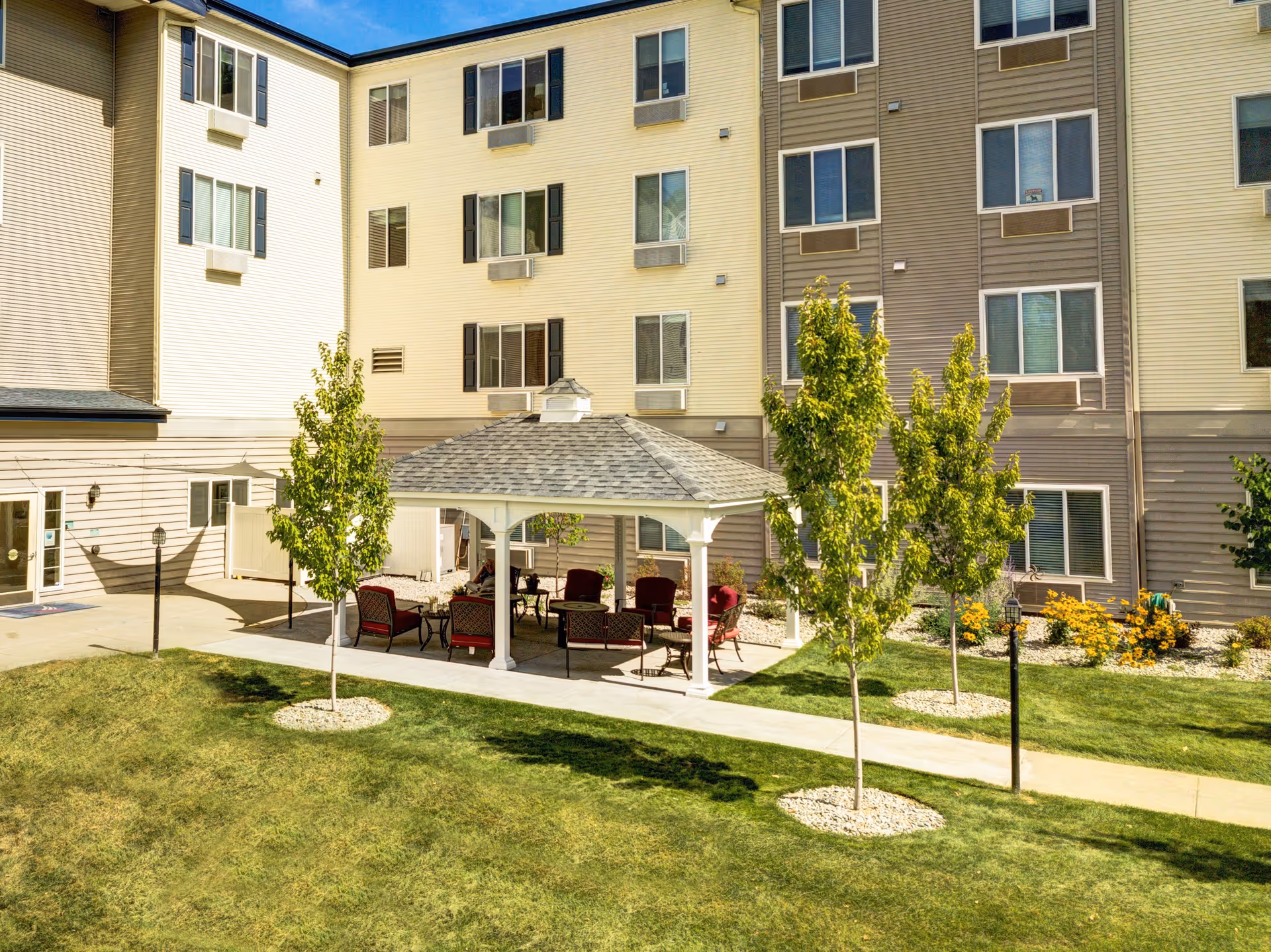 Outdoor courtyard area of a senior living facility with a gazebo featuring several cushioned chairs and tables underneath. The surrounding area has green grass, small trees with mulch around their bases, and a multi-story building with numerous windows in the background.