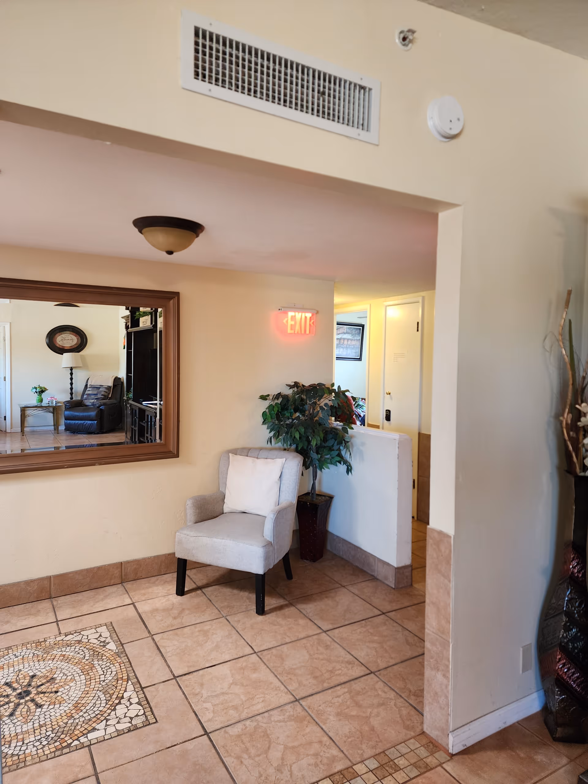 Interior view of a living area in Morning Glory Assisted Living featuring a beige armchair with a white pillow, a potted plant, a large wall mirror reflecting another seating area with a lamp and armchair, tiled floor with mosaic design, and an illuminated exit sign above a doorway.