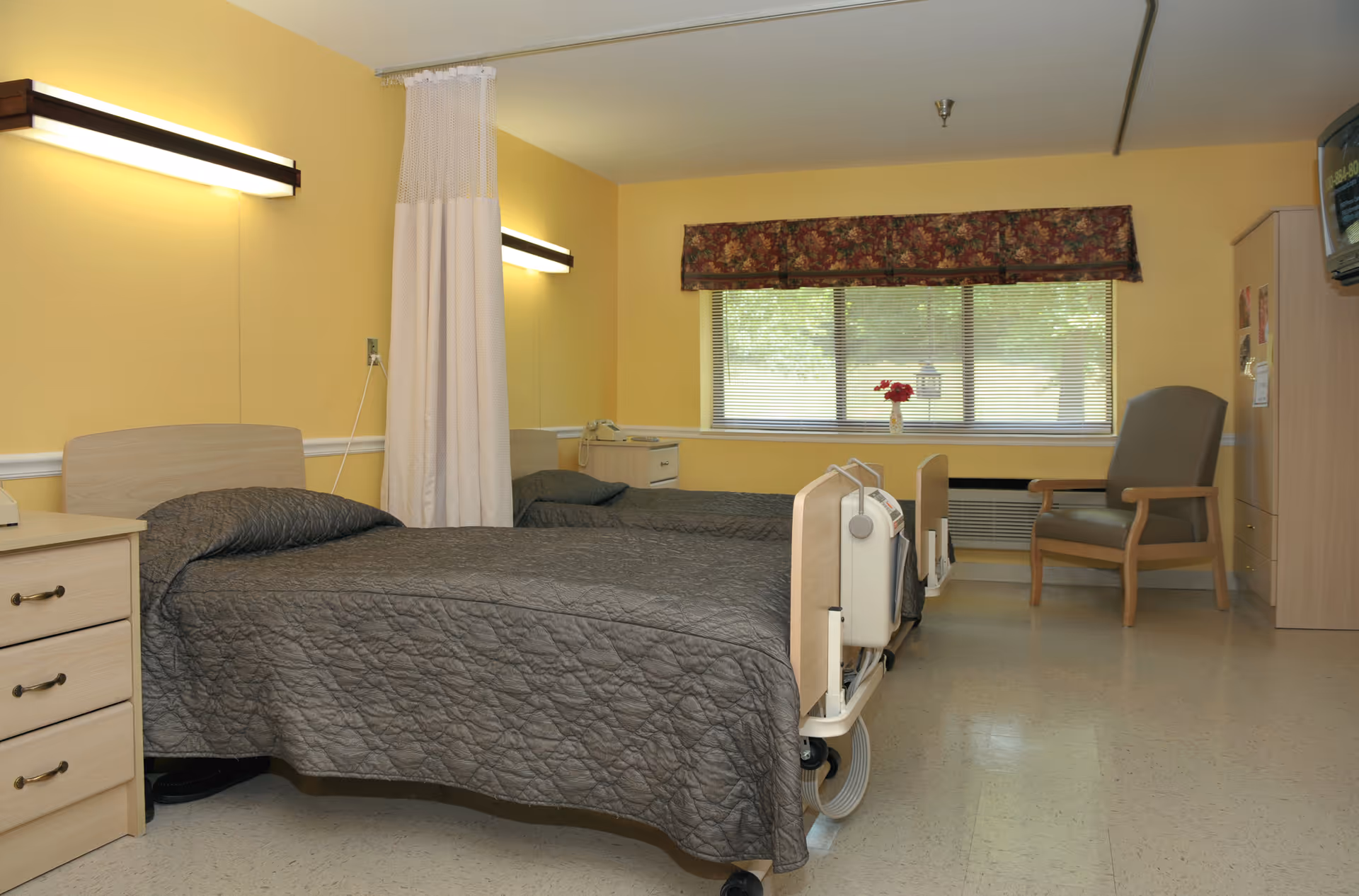 A senior living facility bedroom with two hospital-style beds, a dresser, chair, and a window with a floral valance.