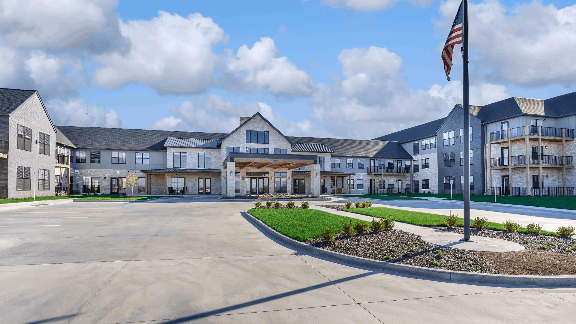 Front exterior view of Cedarhurst Senior Living of Wentzville, showing a large, modern three-story building with multiple windows, balconies, and a covered entrance. The foreground features a paved driveway, landscaped green areas with small bushes, and an American flag on a flagpole. The sky is partly cloudy.