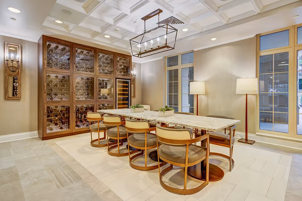 A well-lit dining room featuring a long marble table surrounded by eight wooden chairs with cushioned seats. Behind the table is a large wooden wine rack filled with bottles, and two wall sconces with candles flank the rack. The room has large windows with beige frames, two floor lamps with white shades, and a modern rectangular chandelier hanging from the ceiling.