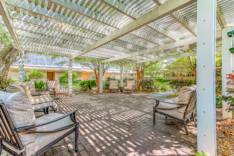 Outdoor seating area at Woodmont Senior Living with cushioned chairs arranged under a white wooden pergola, surrounded by greenery and trees with sunlight casting shadows on the brick floor.