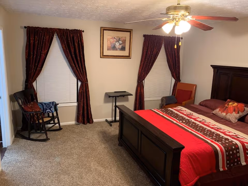 Well-lit bedroom with a dark wood bed dressed in red bedding, a ceiling fan, two curtained windows, a rocking chair and carpeted floor.