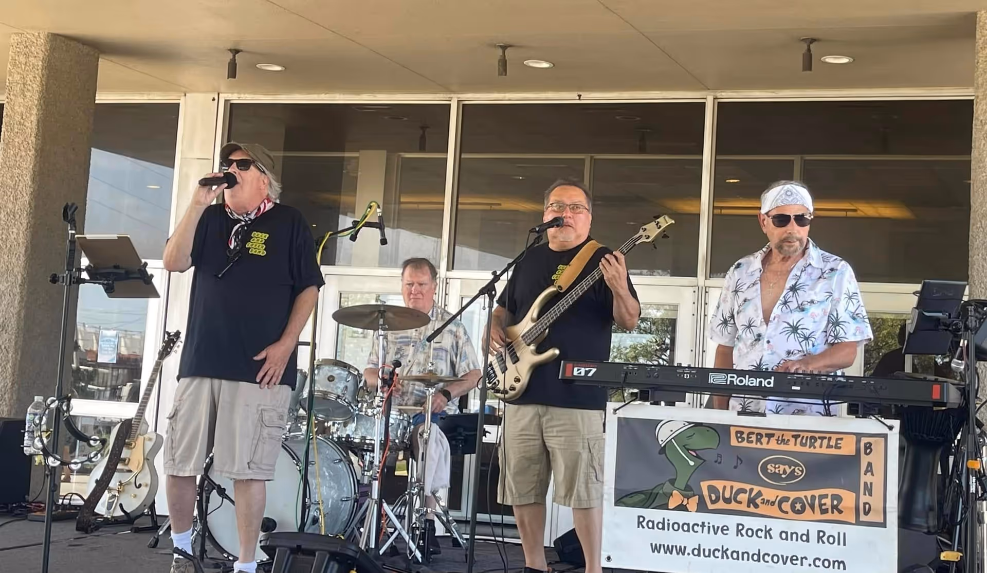 A four-member band performing on an outdoor stage in front of a building. One member is singing into a microphone, another is playing drums, the third is playing bass guitar and singing, and the fourth is playing a keyboard. The keyboard stand has a sign that reads 'Bert the Turtle says Duck and Cover Band, Radioactive Rock and Roll, www.duckandcover.com'.