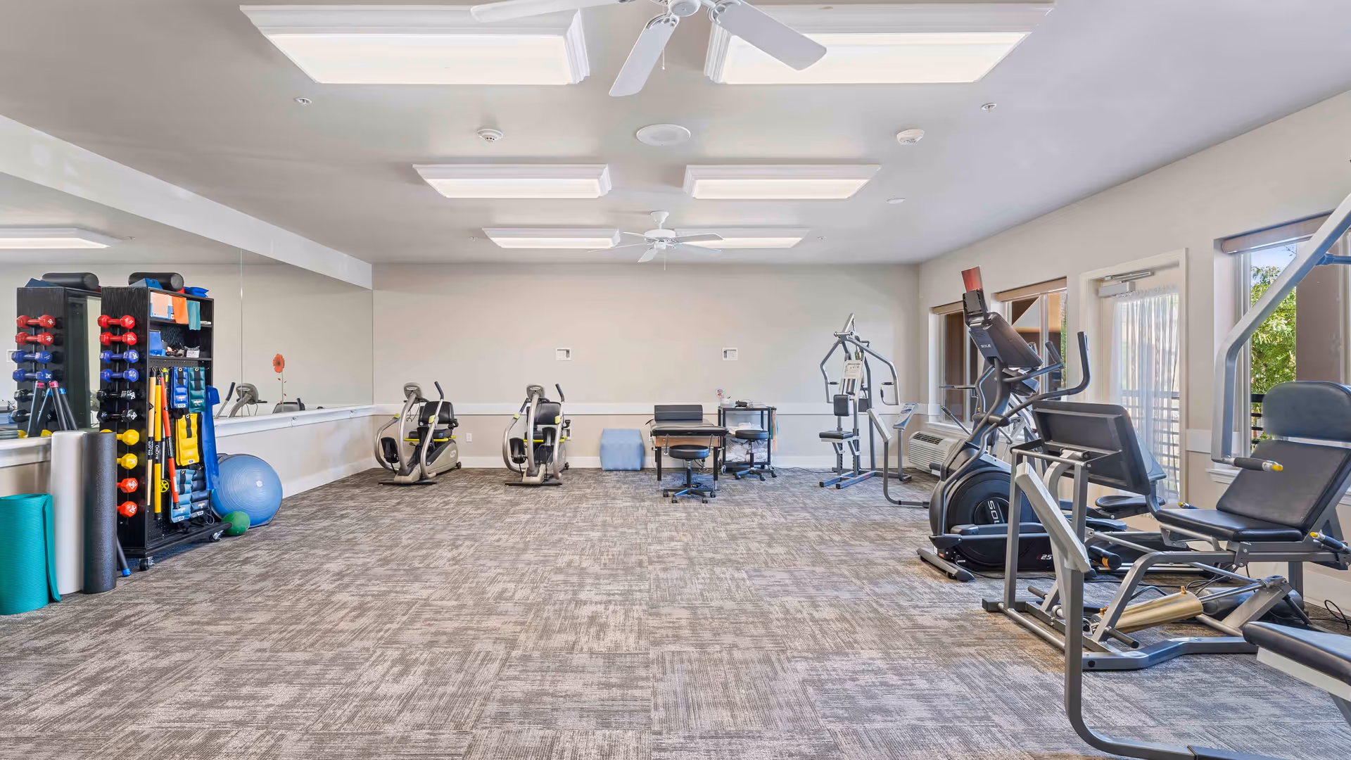 A spacious fitness room with various exercise equipment including stationary bikes, weight machines, free weights, exercise balls, and yoga mats. The room has large windows allowing natural light, ceiling fans, and a mirrored wall on one side.