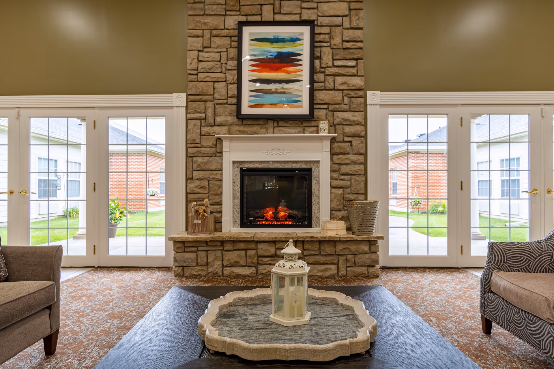 A cozy living room area with a stone fireplace in the center, flanked by large glass doors on both sides that open to a green outdoor courtyard. The fireplace has a colorful abstract painting above it and decorative items on the mantel. Two armchairs are partially visible on either side of the image, and a coffee table with a decorative lantern is in the foreground.