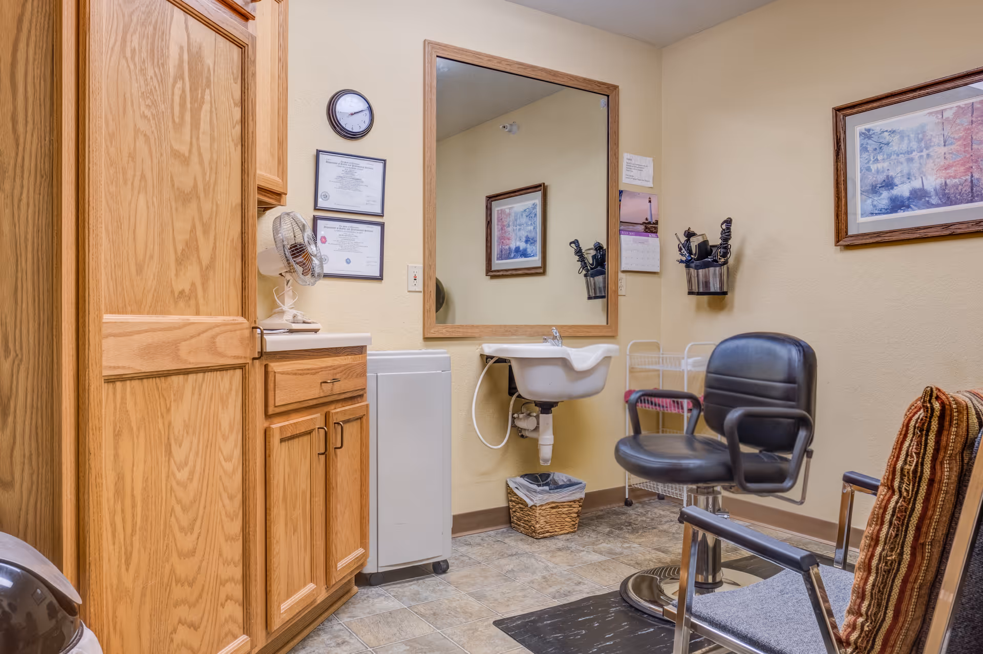 Small hair salon/styling room with a mirror and sink, a salon chair, wooden cabinets and framed artwork on the walls.