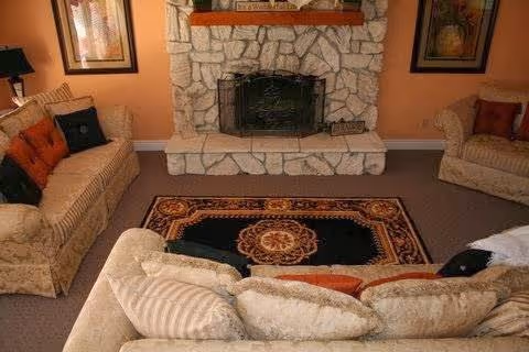 A cozy living room with beige sofas arranged around a decorative area rug featuring a central medallion pattern. The room has a stone fireplace with a black metal screen and a wooden mantel. Two framed artworks hang on the peach-colored walls on either side of the fireplace.