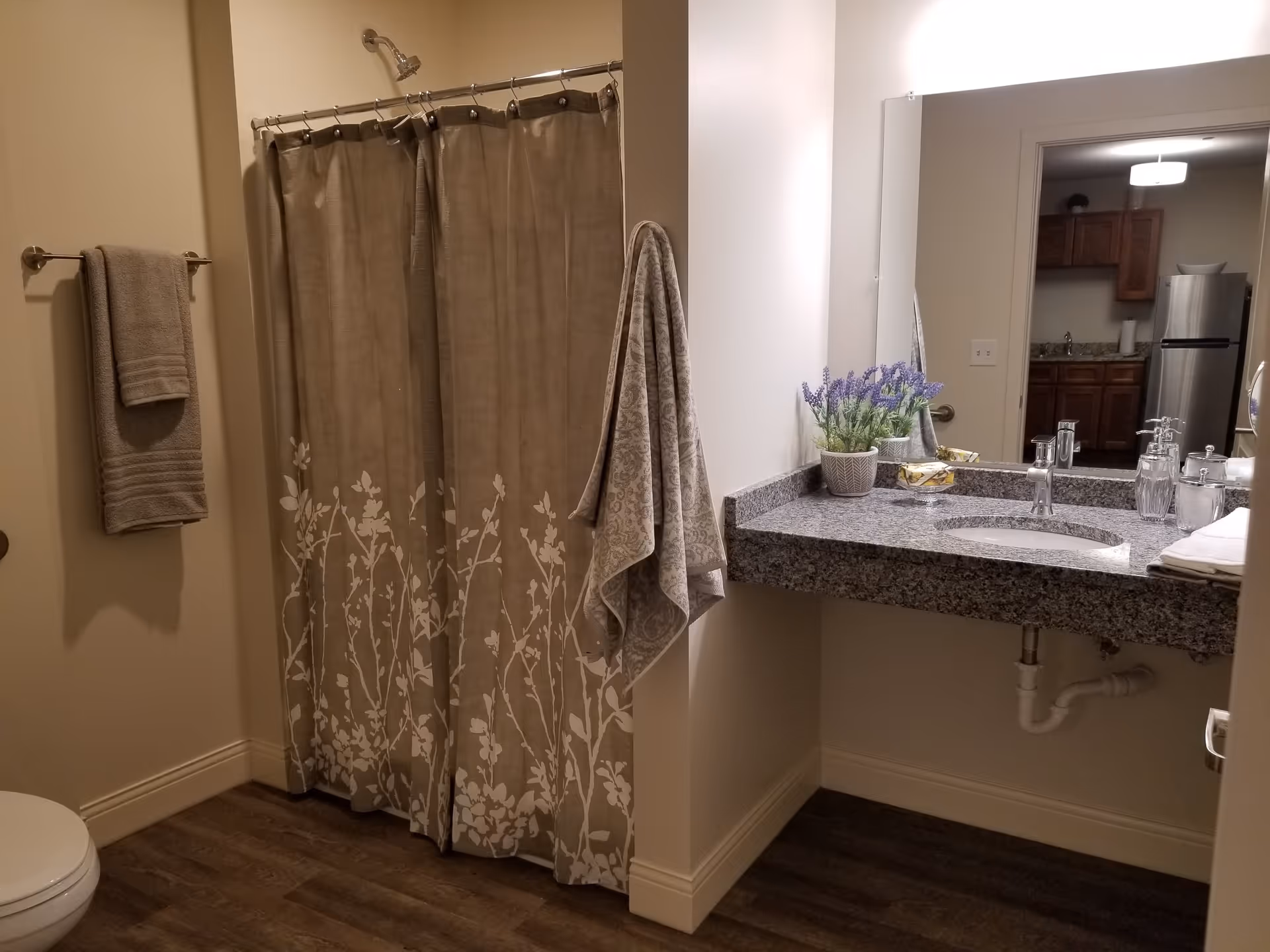 A bathroom with a shower area covered by a beige curtain with white floral patterns. There are two beige towels hanging on the walls, a granite countertop with a sink, a potted plant with purple flowers, soap, and other toiletries. A large mirror above the sink reflects a kitchen area with wooden cabinets and a stainless steel refrigerator.