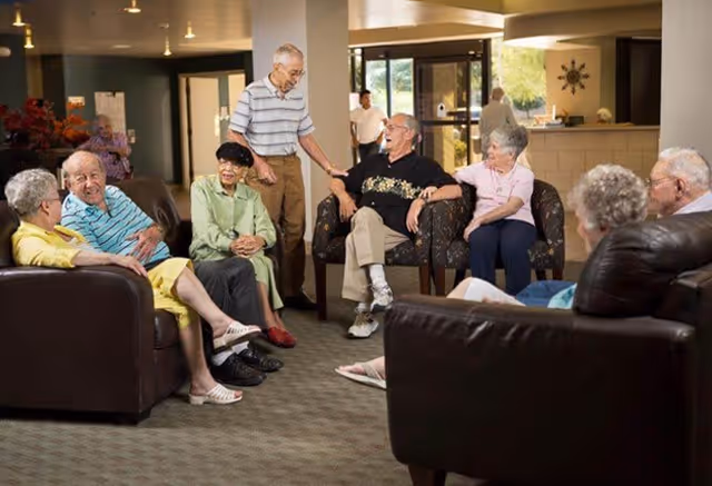 A group of elderly people sitting and standing in a spacious, well-lit common area of a senior living facility, engaging in conversation and smiling. The room has comfortable chairs and sofas arranged in a circle, with a reception desk visible in the background.