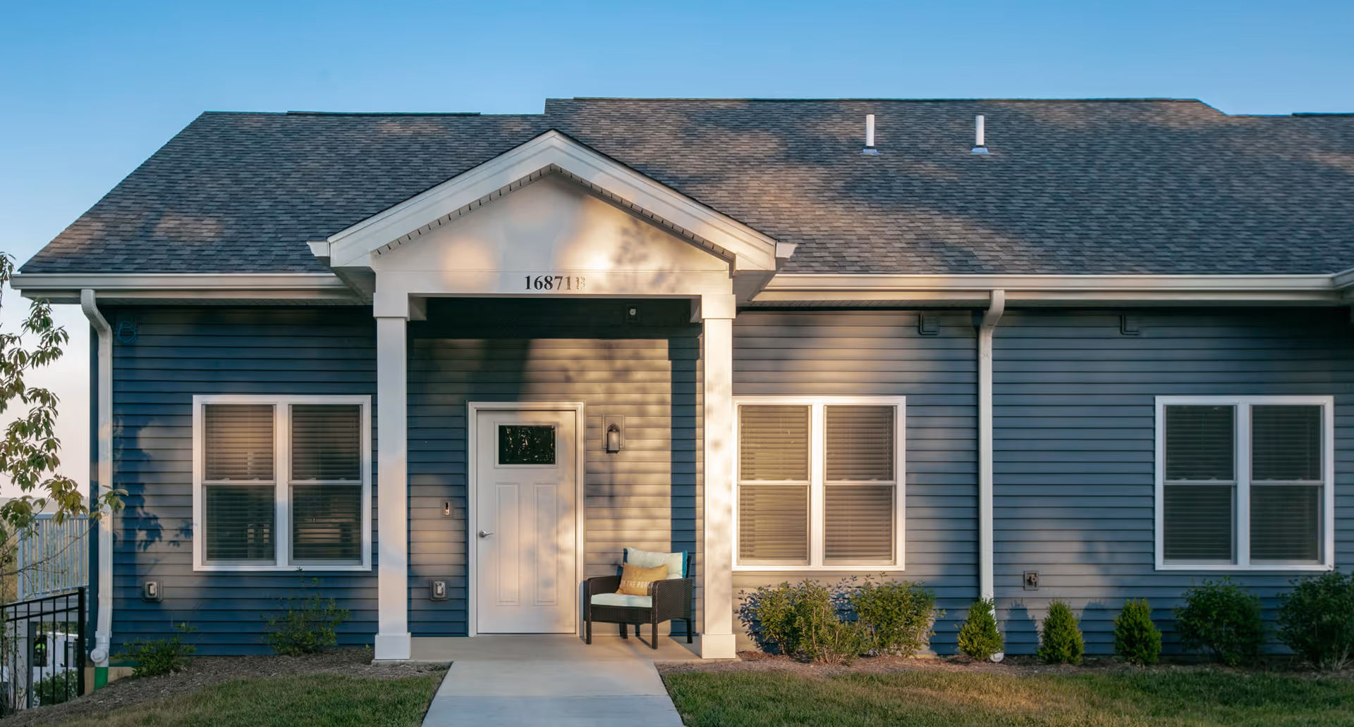 Front exterior view of a single-story building with blue siding, white trim, and a gray shingled roof. The entrance features a small covered porch with two white columns, a white door with a small window, and a chair with cushions. There are three sets of windows with closed blinds and some landscaping with bushes and grass in front.