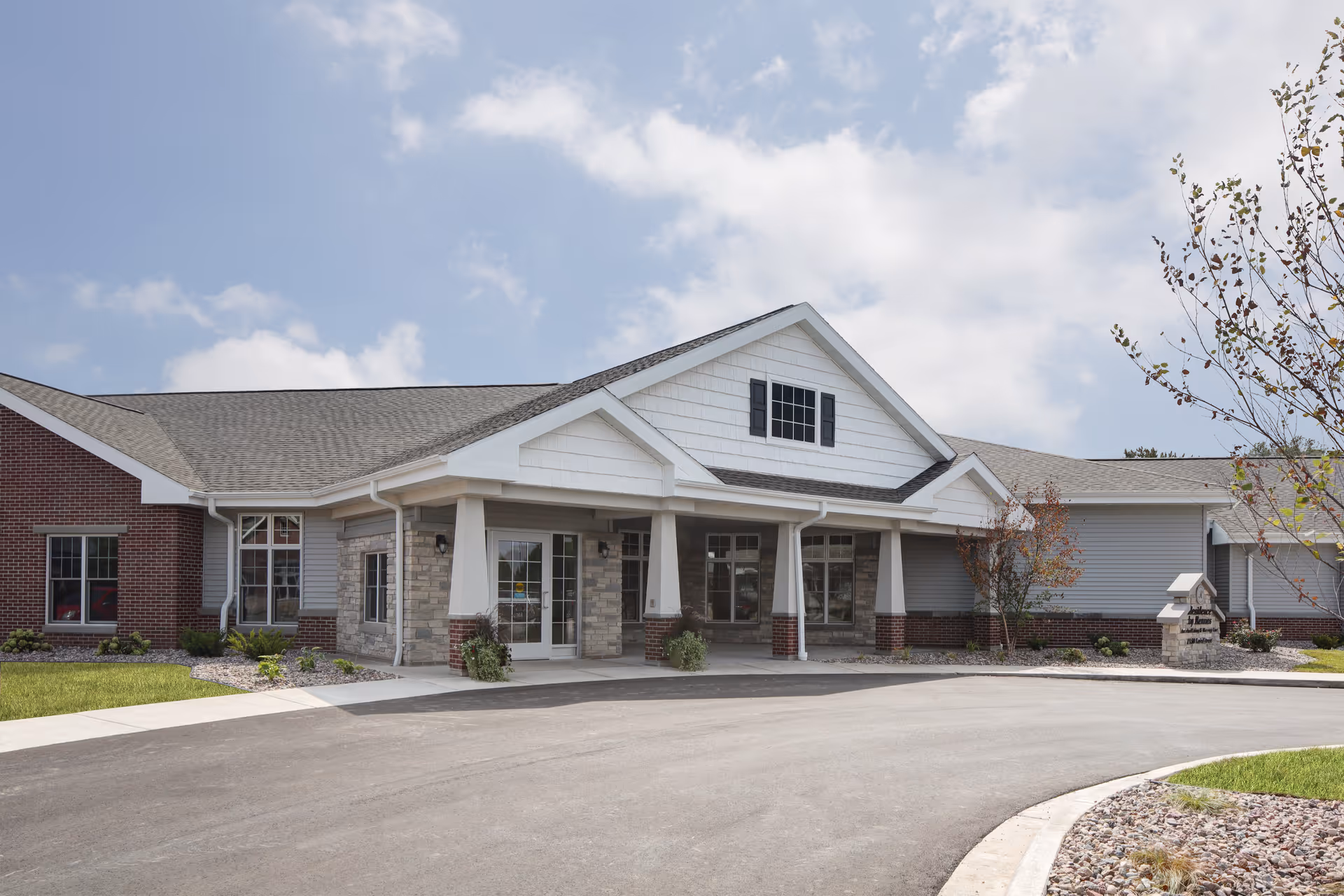 Front exterior view of a single-story assisted living and memory care facility with a covered entrance, brick and siding exterior, and a driveway leading up to the entrance. There are small landscaped areas with grass, rocks, and young trees around the building.
