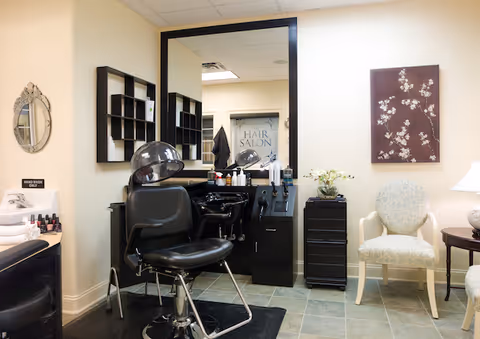 Small salon room with a black styling chair and hooded dryer facing a large mirror, shelving, and a decorative seating area.