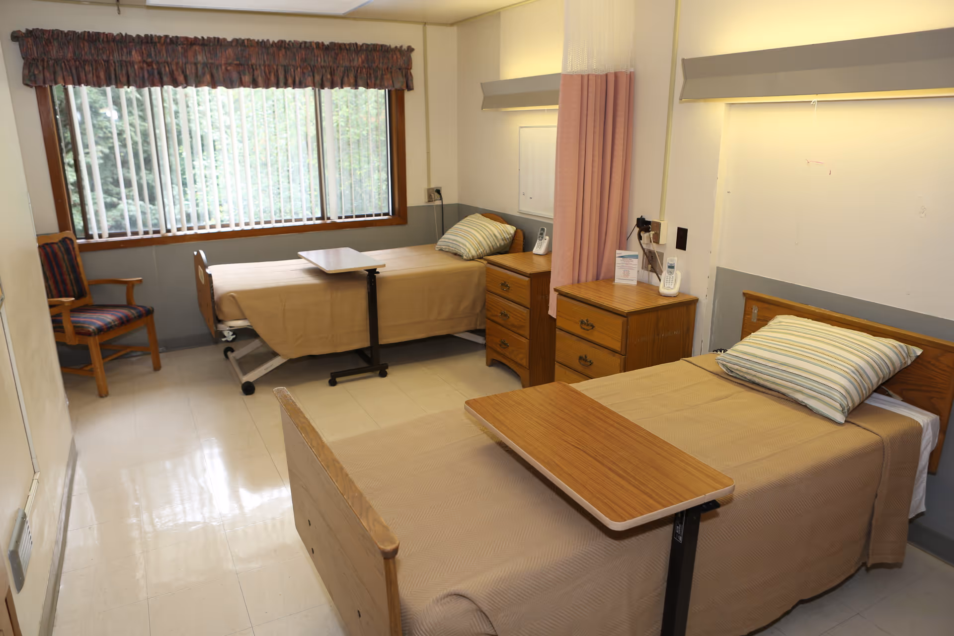 A room with two single hospital-style beds each with a beige blanket and a striped pillow. Each bed has a wooden overbed table. There are two wooden nightstands between the beds, a wooden chair with a striped cushion near a large window with vertical blinds and a valance. The room has a light-colored tiled floor and pale walls with a pink privacy curtain between the beds.
