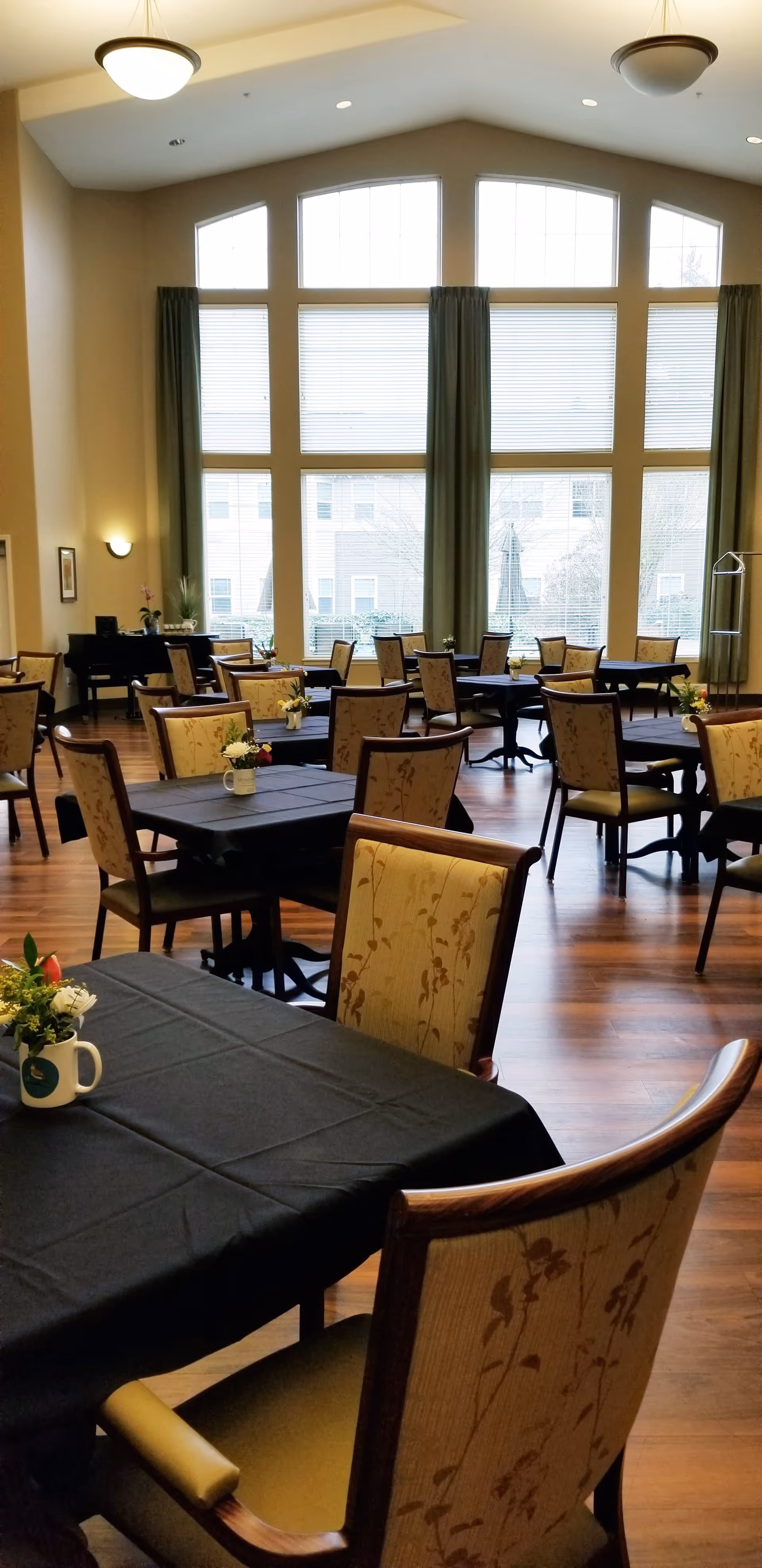 A spacious dining room with multiple tables covered in black tablecloths and surrounded by upholstered chairs with floral patterns. Large windows with green curtains allow natural light to fill the room, and small flower arrangements are placed on each table. The room has wooden flooring and high ceilings with hanging light fixtures.