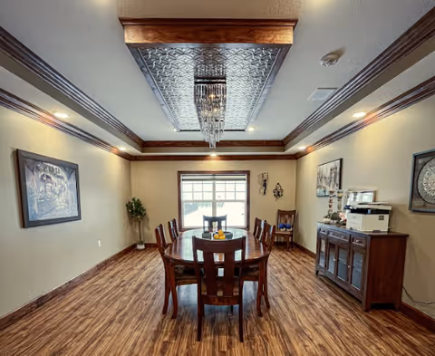 A communal dining room with a wooden table and chairs beneath a decorative ceiling panel and chandelier, hardwood floors and a sideboard.
