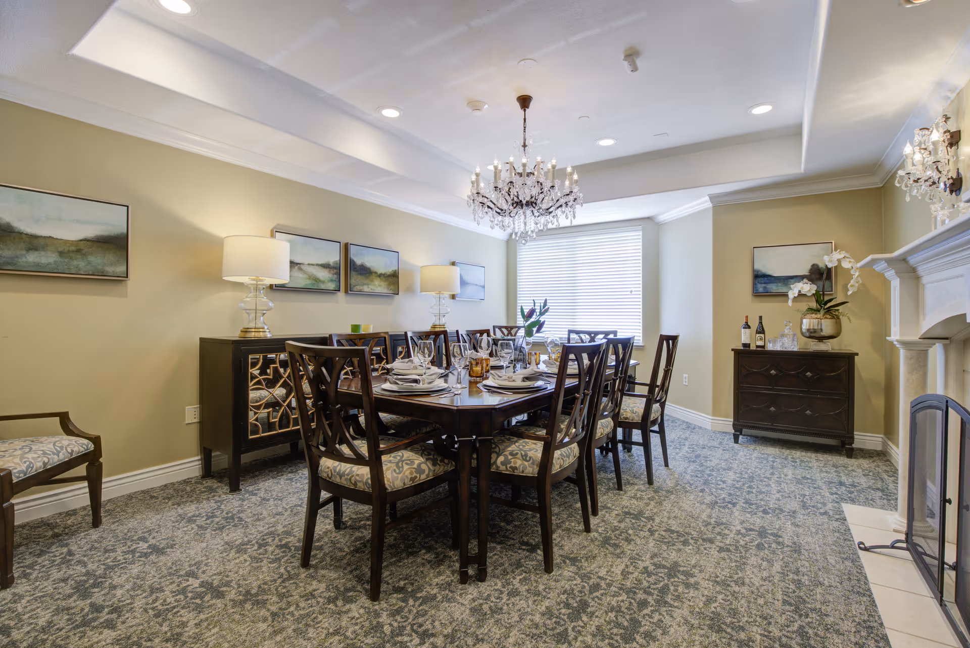 A formal dining room with a large wooden dining table set for eight people. The chairs have patterned cushions. The room features a chandelier hanging from the ceiling, two table lamps on sideboards, framed landscape paintings on the walls, a window with blinds, and a fireplace with a decorative screen. The carpet has a floral pattern.