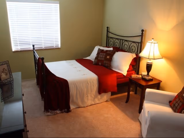 A cozy bedroom with a metal-framed bed dressed in white and red bedding, a bedside table with a lamp, a white armchair with a decorative pillow, and a window with closed blinds letting in natural light.