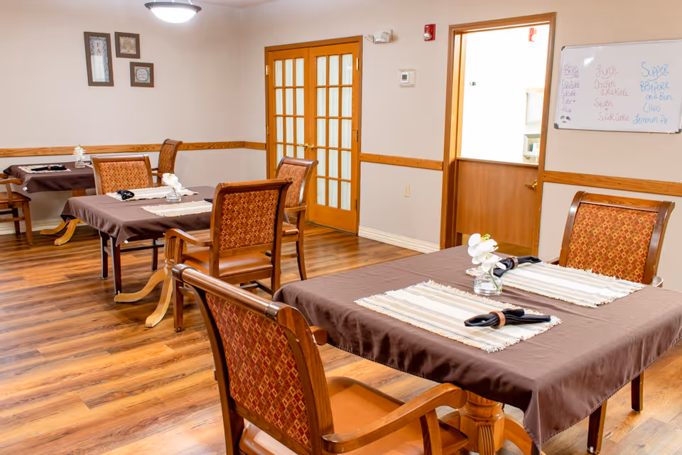 A dining room with wooden floors and several tables covered with brown tablecloths and beige placemats. Each table has wooden chairs with patterned upholstery and a small flower vase centerpiece. There are framed pictures on the wall, a whiteboard with handwritten notes, and a set of wooden double doors with glass panels.