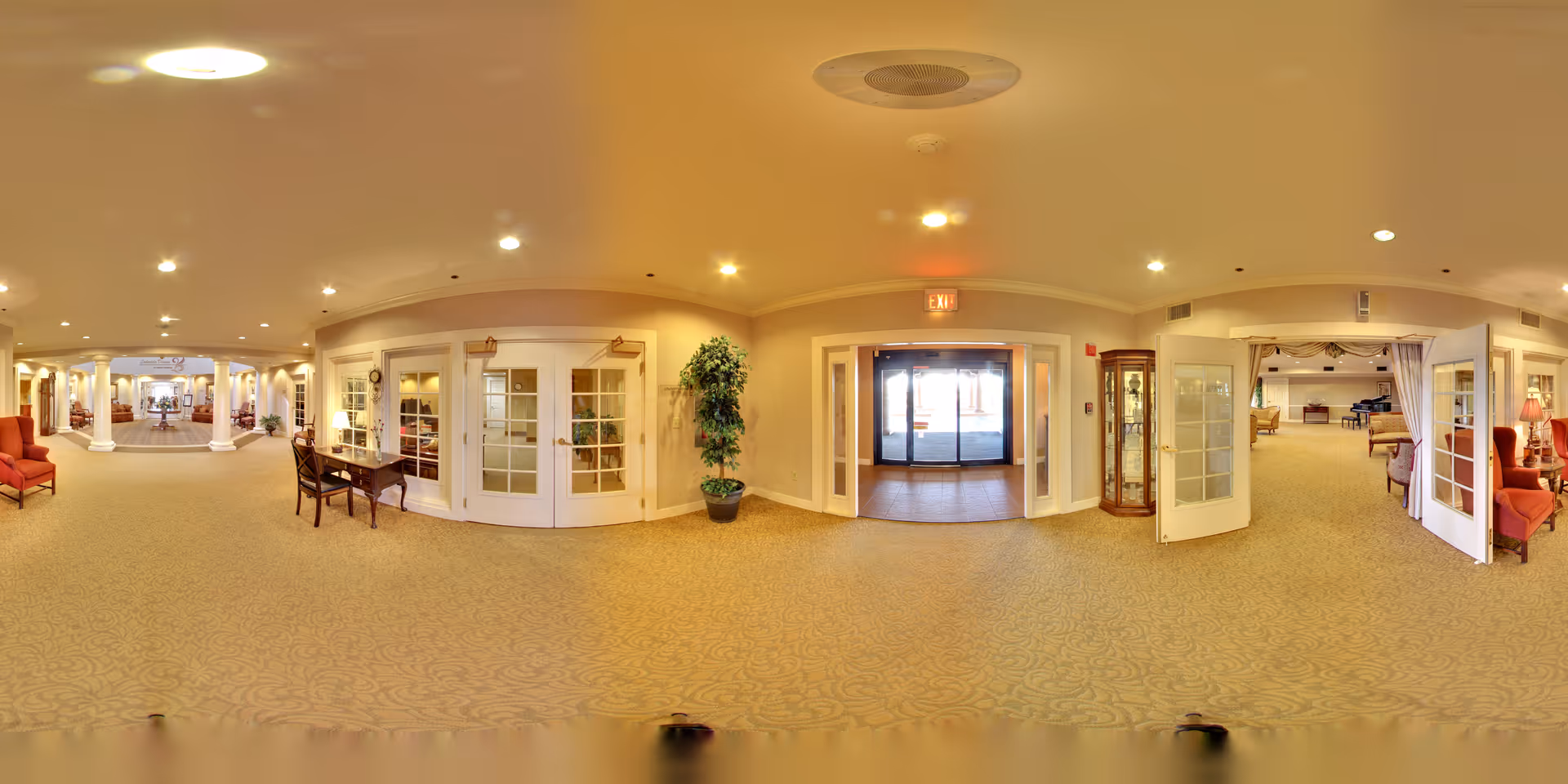 Wide panoramic view of a warmly lit senior living facility lobby with carpeted floors, seating areas, columns, and glass entrance doors.