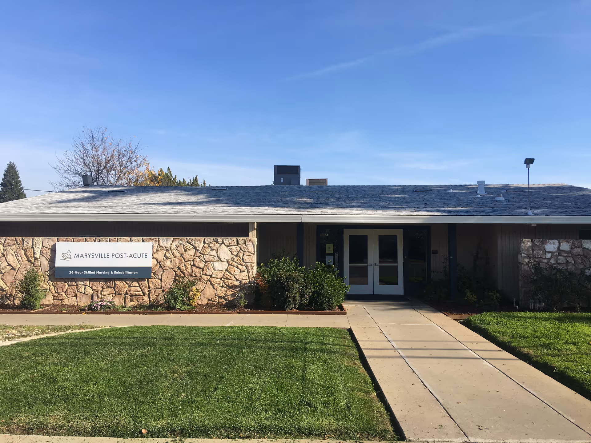Front exterior view of Marysville Post-Acute facility showing a single-story building with stone and wood facade, a sign with the facility name and description, a concrete walkway leading to double glass doors, green lawn, and clear blue sky.