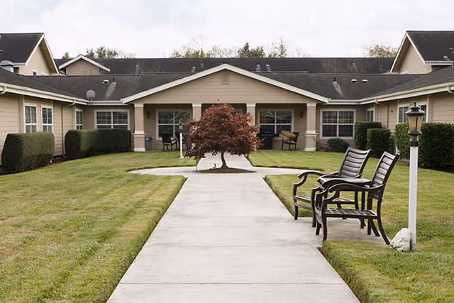 A courtyard area of a senior living community with a concrete walkway leading to a single-story building. The walkway is flanked by green grass and trimmed bushes. There is a small tree in the center of the courtyard and two benches with armrests on the right side near a lamp post.