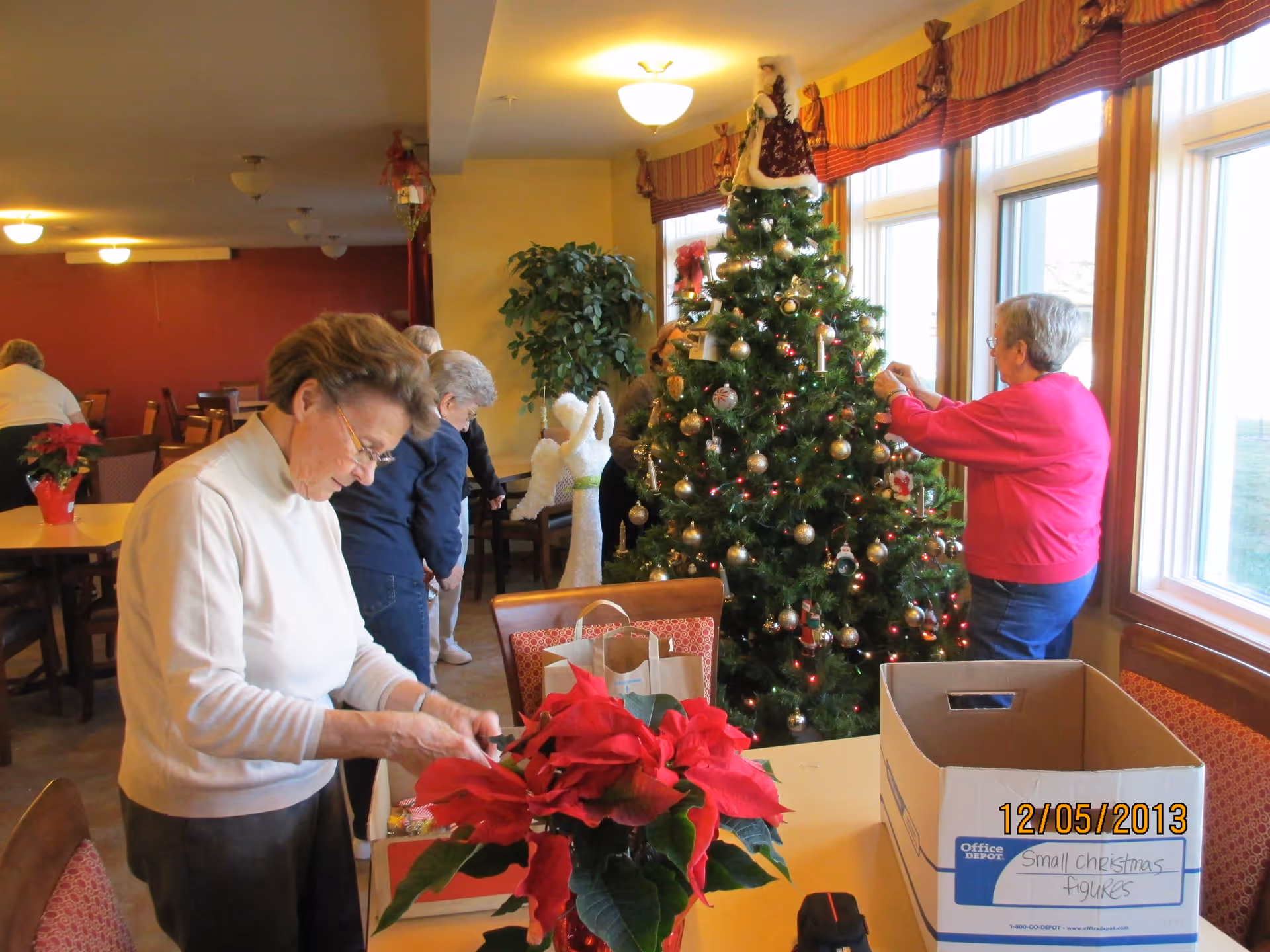 Residents decorate a Christmas tree in a bright dining/common room with tables and poinsettias.