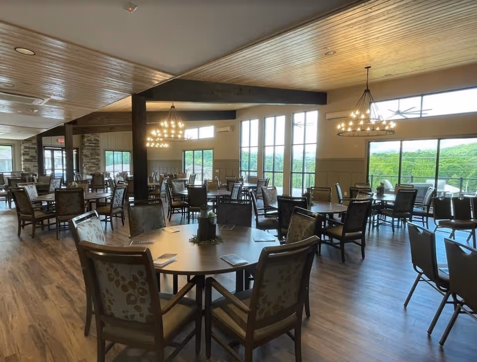 Spacious dining room with multiple round tables and cushioned chairs arranged neatly. Large windows and glass doors allow natural light to fill the room, offering a view of greenery outside. Two large chandeliers hang from the wooden ceiling, and the floor is covered with wood-style flooring.