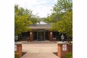 Entrance to Lemont Nursing and Rehabilitation Center with a covered walkway, flanked by brick pillars and large planters, surrounded by green trees and shrubs.