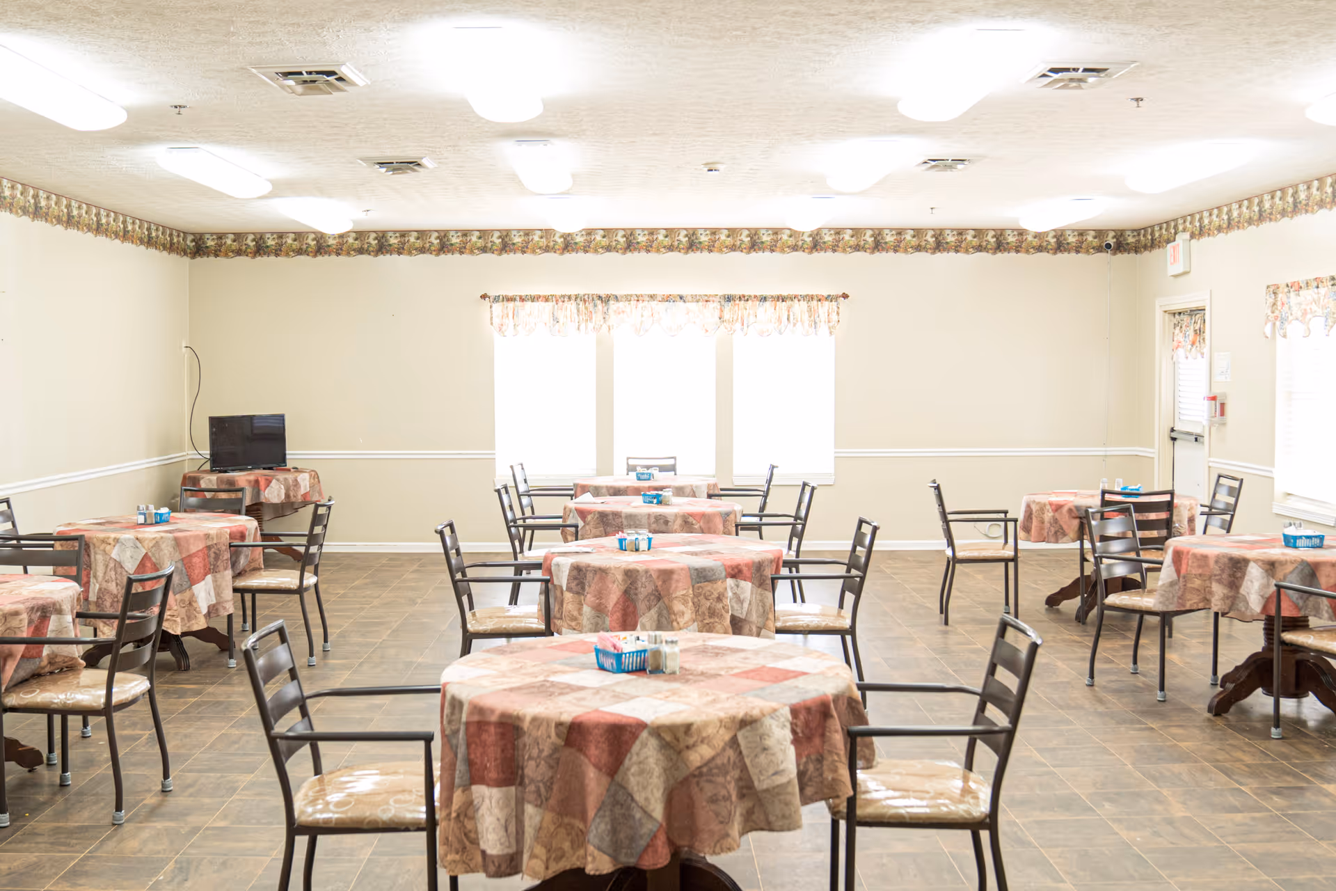 A bright dining room with multiple round tables covered in patterned tablecloths. Each table is surrounded by metal chairs with cushioned seats. The room has tiled flooring, beige walls with a floral border near the ceiling, and several windows with floral valances allowing natural light to enter. A small TV is placed on a table in the corner.