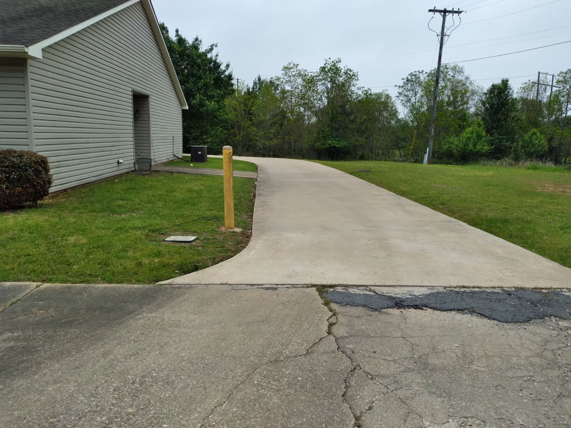 A concrete driveway leading away from a building with beige siding. The driveway is bordered by green grass and bushes, with trees and utility poles visible in the background under a cloudy sky.