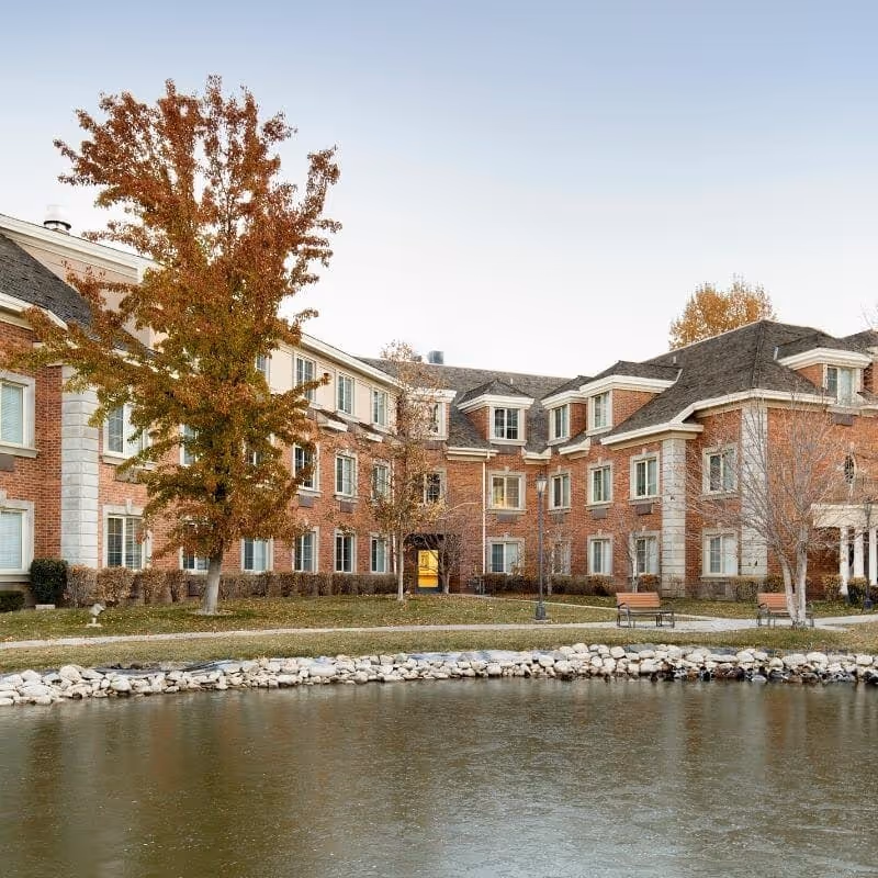Exterior view of a multi-story brick senior living facility named Courtyard at Jamestown, with several windows, a few trees with autumn foliage, benches, a walking path, and a pond in the foreground.