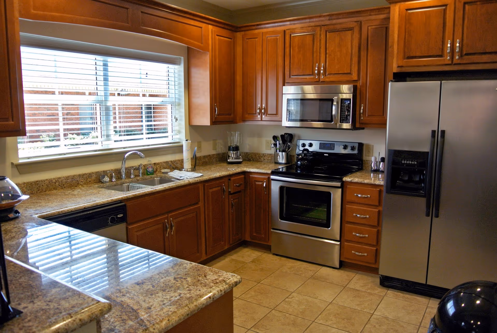 A modern kitchen with wooden cabinets, granite countertops, a stainless steel refrigerator, oven, microwave, and a double sink under a window with white blinds.