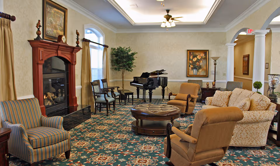 A cozy living room area with patterned carpet, a grand piano, a fireplace with a wooden mantel, several upholstered chairs and a sofa, a round leather ottoman, framed artwork on the walls, and a potted plant near a window with beige curtains.