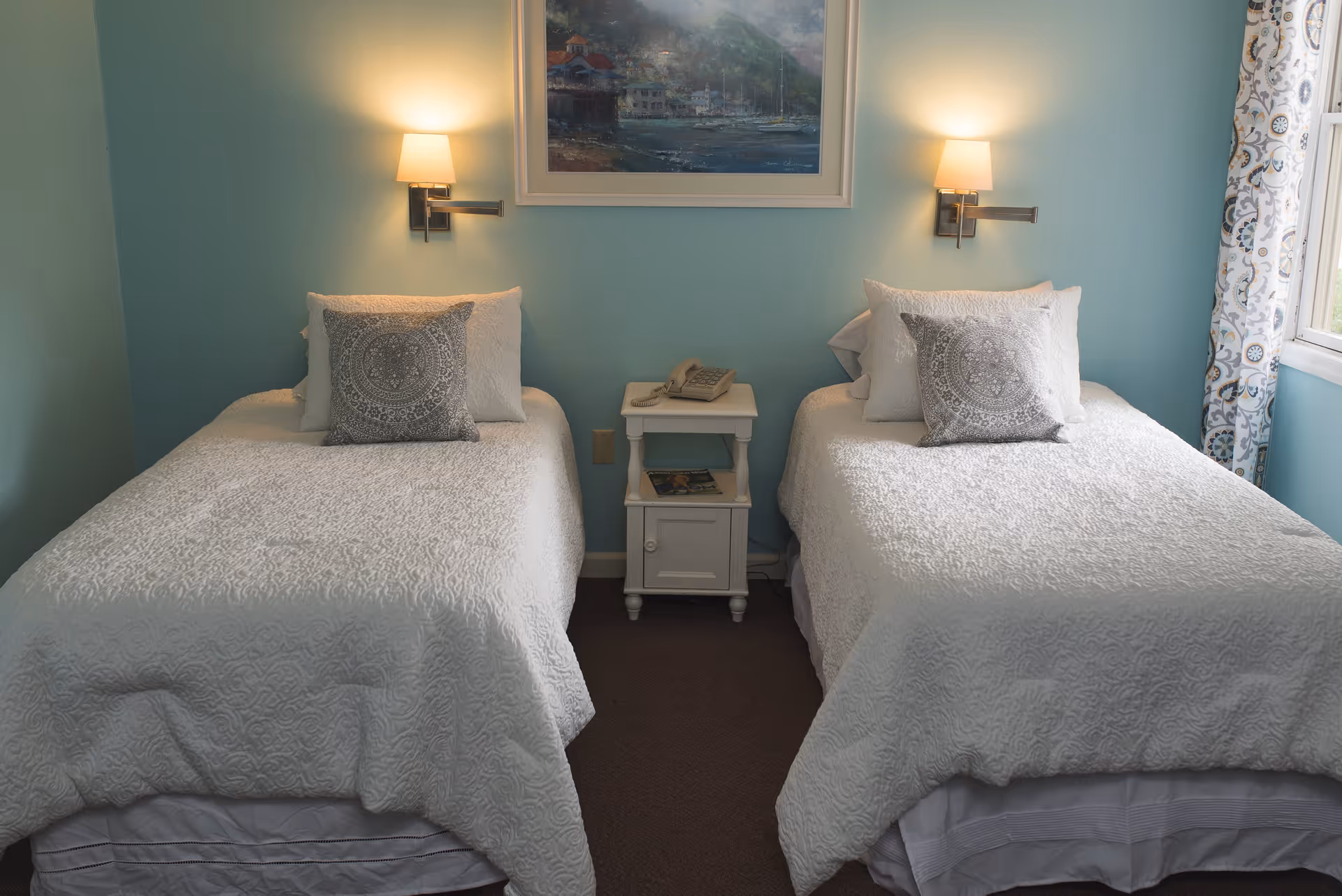 A bedroom with two twin beds, each with white bedding and a decorative pillow. A small white nightstand with a telephone and a magazine sits between the beds. Two wall-mounted lamps are above each bed, and a framed painting hangs on the light blue wall. A window with patterned curtains is visible on the right side.