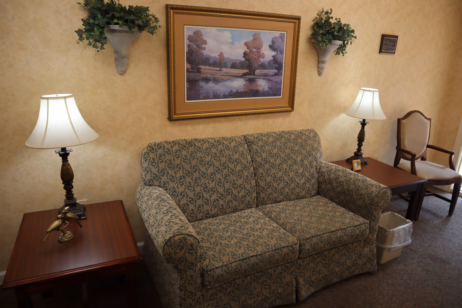 A cozy living room area with a patterned sofa, two wooden side tables each with a lamp, a framed landscape painting on the wall, two wall-mounted plant holders with greenery, an upholstered armchair, and a small trash bin on the carpeted floor.