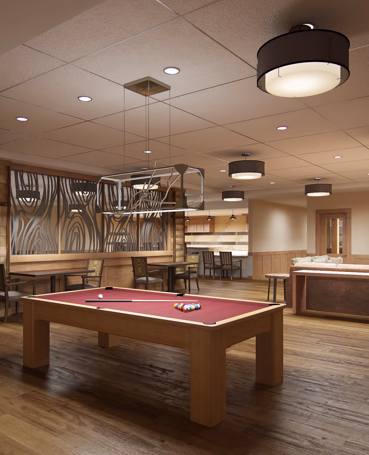 Interior view of a senior living facility common area featuring a pool table with a red felt top and pool cues, several wooden tables and chairs, a seating area with a sofa, and a kitchen area in the background. The room has wooden flooring, decorative wall panels, and modern ceiling lights.