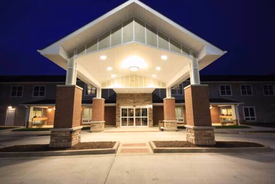 Entrance of a senior living facility at night with a well-lit covered drop-off area supported by brick and stone pillars, and glass double doors leading inside.
