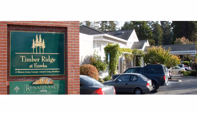Exterior view of Timber Ridge at Eureka assisted living residence showing a green sign with the facility name mounted on a brick pillar, and a parking lot with several cars parked in front of white buildings surrounded by trees and shrubs.