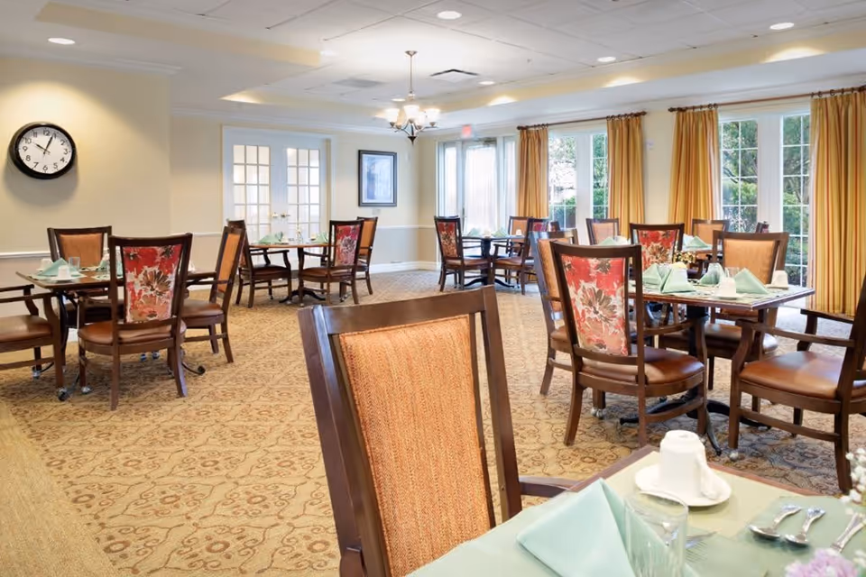 A bright dining room with multiple wooden tables and chairs, each table set with green napkins, white cups, and silverware. The room has large windows with yellow curtains allowing natural light to enter, a wall clock, and a chandelier hanging from the ceiling.