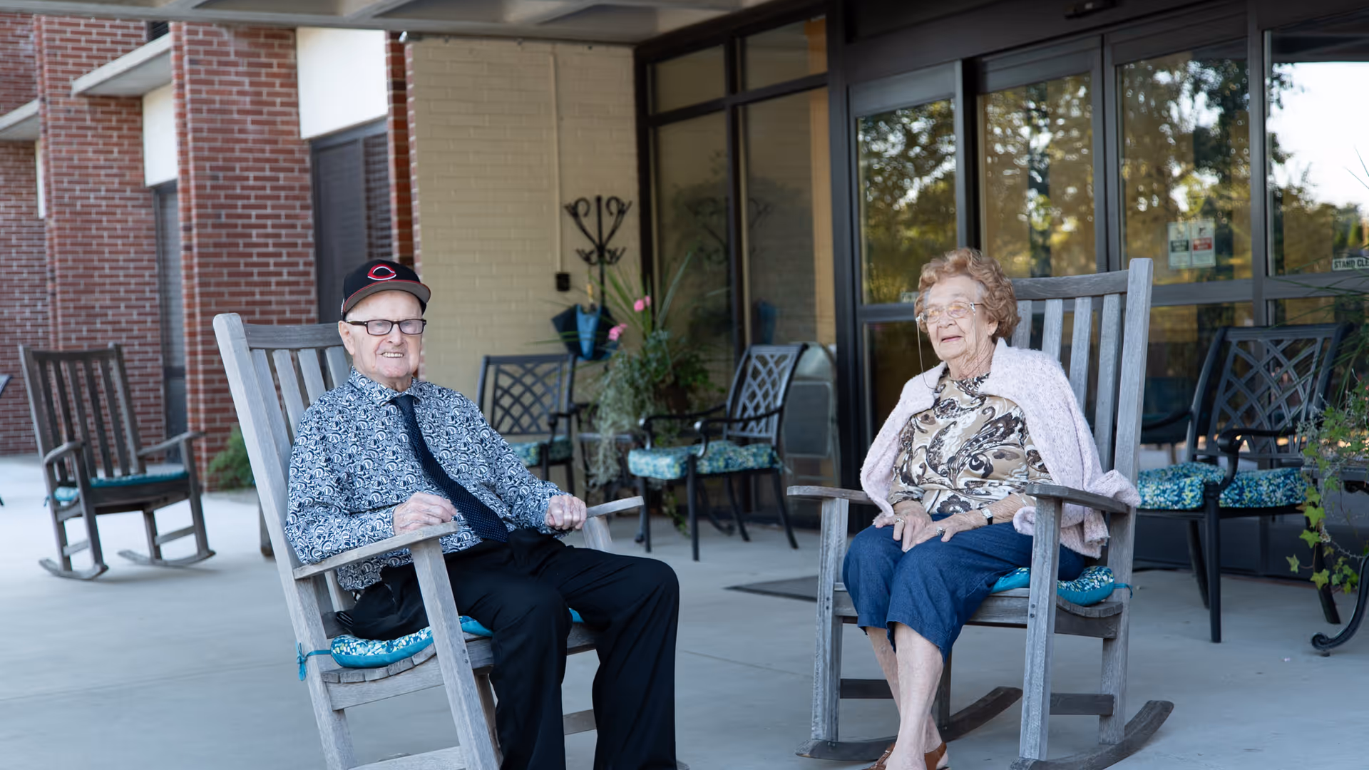 An elderly man and woman sitting on wooden rocking chairs on a covered patio outside a building. The man is wearing a patterned shirt, black pants, a tie, and a baseball cap, while the woman is wearing a patterned blouse, blue pants, and a light pink shawl. Behind them are more chairs and a glass door entrance with reflections of trees.