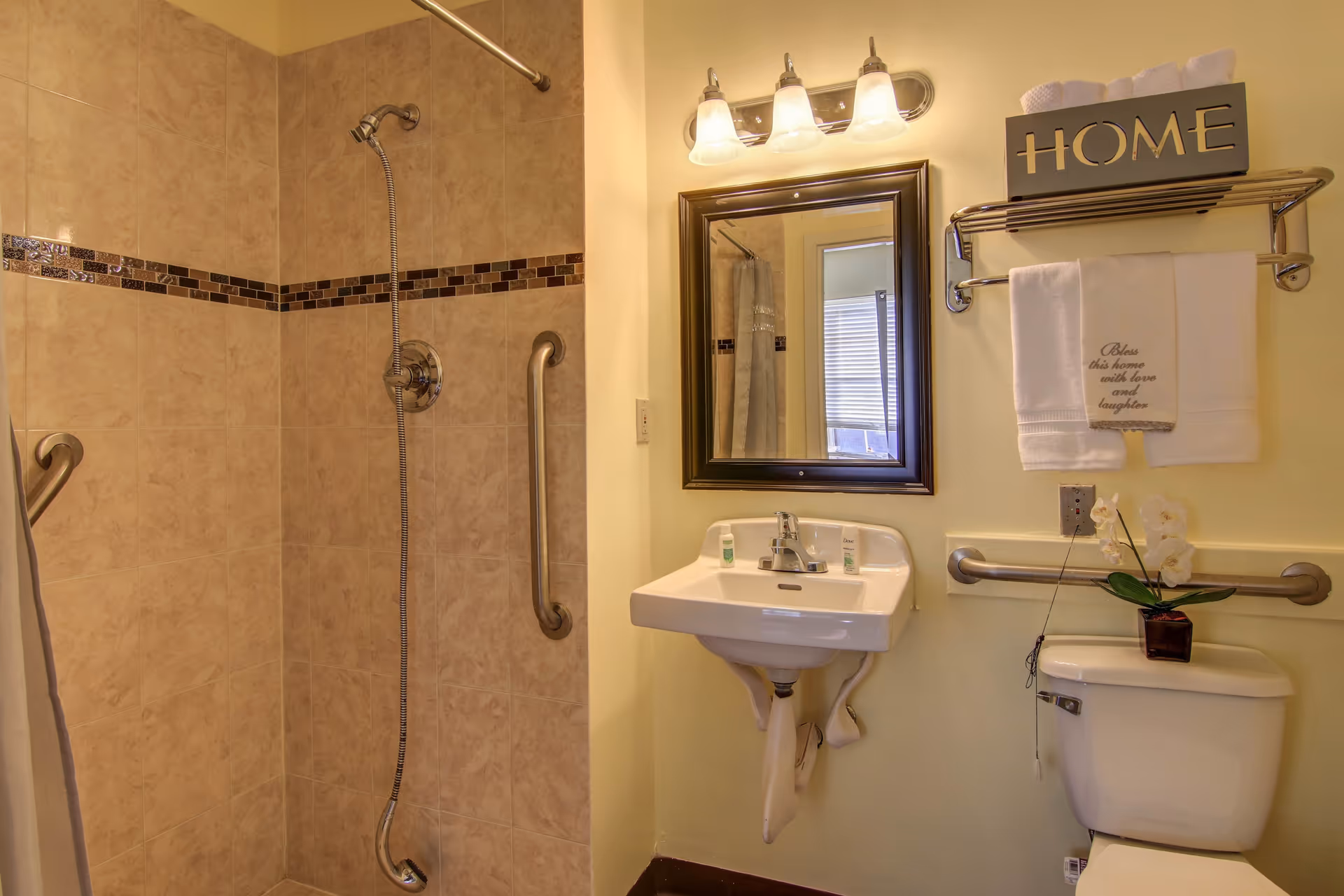 Accessible bathroom with a tiled walk-in shower, grab bars, wall-mounted sink and mirror, towel rack and toilet.