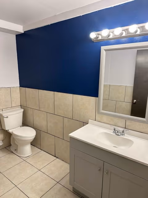 A bathroom with beige tiled walls and floor, a white toilet, a white vanity with a sink, a mirror above the sink, and a light fixture with multiple bulbs above the mirror. The upper part of the wall is painted dark blue.