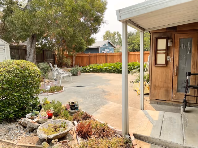 Outdoor patio area with a concrete ground, surrounded by a wooden fence and greenery including bushes and trees. There are two white chairs placed near the fence, a small garden bed with various plants and a stone birdbath in the foreground. On the right side, there is a covered porch attached to a wooden building with a door and window, and a walker is visible near the entrance.